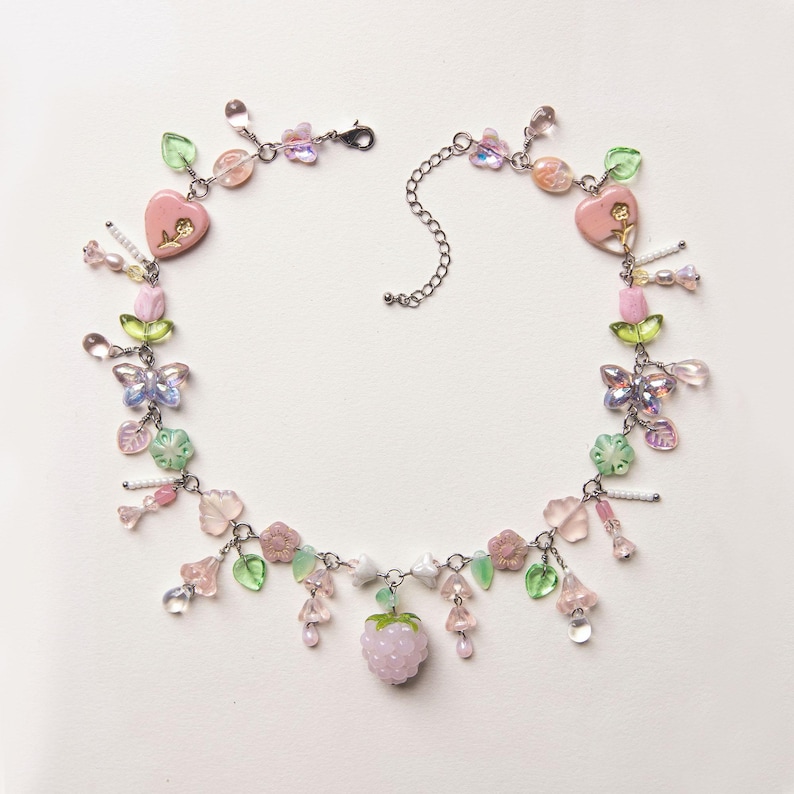 May include: A delicate beaded necklace featuring a variety of pastel-colored beads. The necklace includes pink heart-shaped charms, butterfly and flower accents, and a strawberry pendant. The necklace is made with a silver-colored chain and clasp.