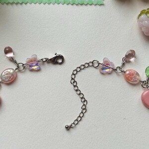 May include: A delicate necklace with pink and iridescent beads, butterfly charms, and a silver chain. The necklace is partially unclasped, showcasing its design. The background includes a green and white checkered fabric and decorative pink berry-shaped beads.