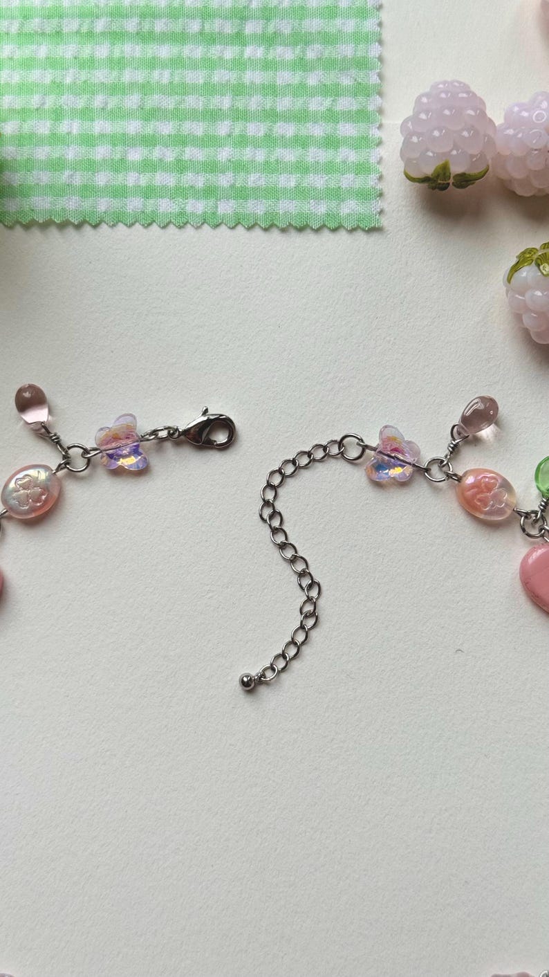 May include: A delicate necklace with pink and iridescent beads, butterfly charms, and a silver chain. The necklace is partially unclasped, showcasing its design. The background includes a green and white checkered fabric and decorative pink berry-shaped beads.