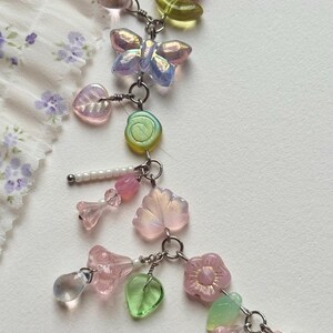 May include: A delicate beaded necklace featuring a variety of pastel-colored charms. The necklace includes butterfly, flower, and leaf-shaped beads, along with heart-shaped pendants and small pearls. The beads are strung on a silver chain.