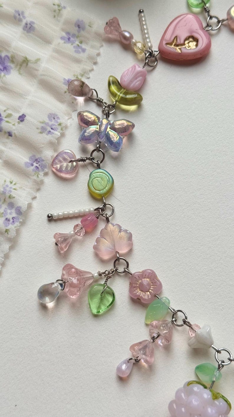 May include: A delicate beaded necklace featuring a variety of pastel-colored charms. The necklace includes butterfly, flower, and leaf-shaped beads, along with heart-shaped pendants and small pearls. The beads are strung on a silver chain.
