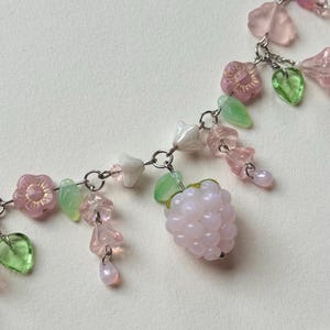 May include: A delicate necklace featuring a silver chain adorned with pink flower and leaf charms, along with a cluster of pink berry beads. The necklace also includes translucent green and pink beads, creating a whimsical and feminine design.
