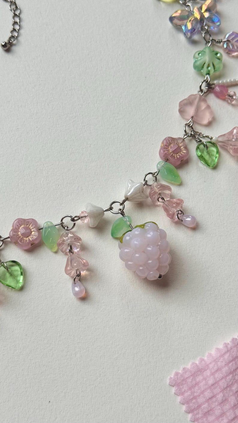 May include: A delicate necklace featuring a silver chain adorned with pink flower and leaf charms, along with a cluster of pink berry beads. The necklace also includes translucent green and pink beads, creating a whimsical and feminine design.
