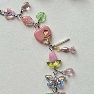 May include: A delicate necklace featuring a variety of colorful beads and charms. The necklace includes pink heart-shaped pendants, iridescent butterfly charms, and small flower and leaf accents. The design incorporates a mix of pink, green, and iridescent elements.