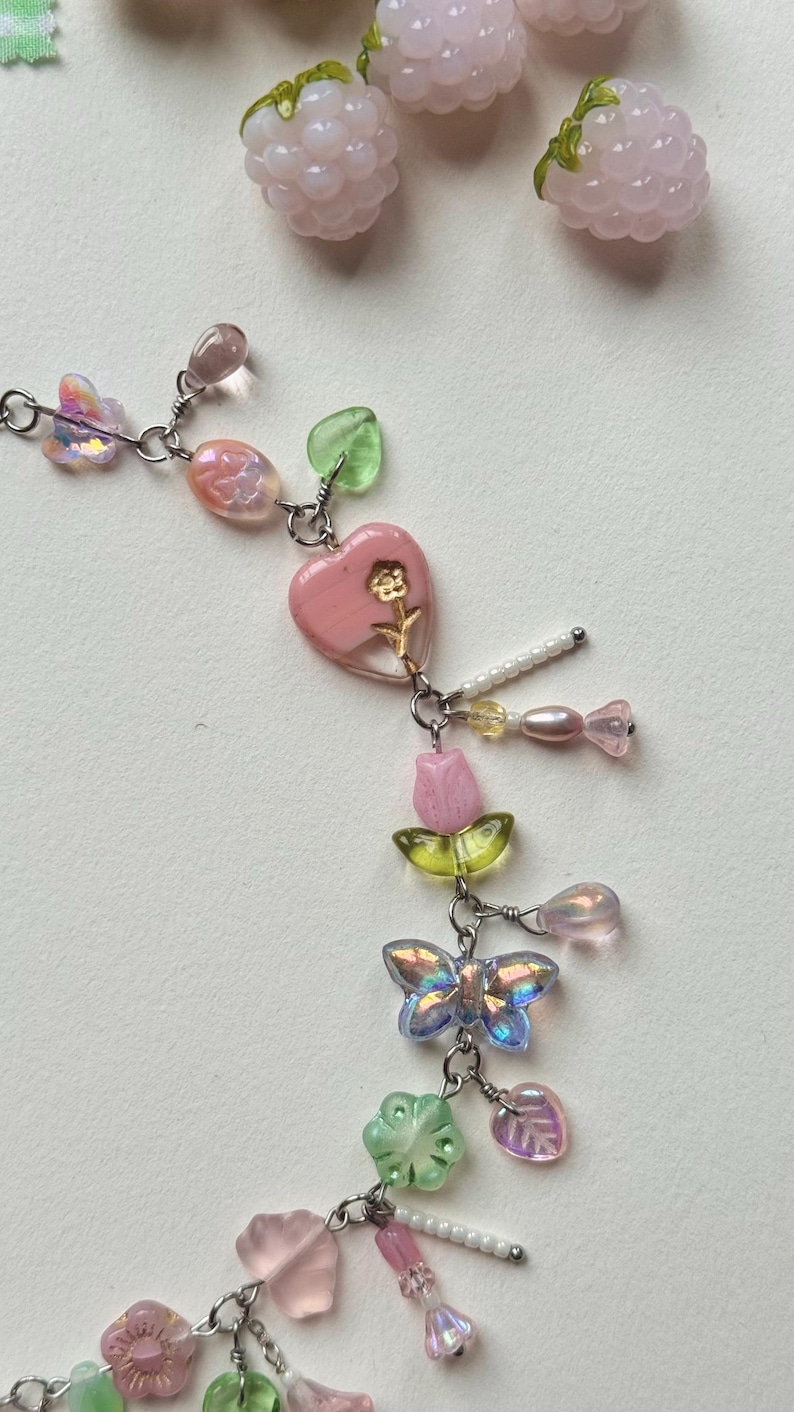 May include: A delicate necklace featuring a variety of colorful beads and charms. The necklace includes pink heart-shaped pendants, iridescent butterfly charms, and small flower and leaf accents. The design incorporates a mix of pink, green, and iridescent elements.