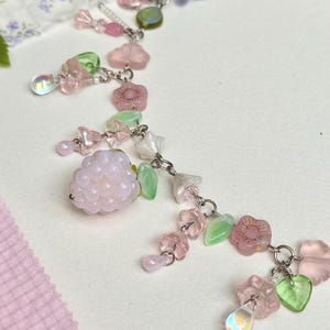 May include: A delicate necklace featuring a variety of pastel-colored beads and charms. The necklace includes pink, green, and clear beads, along with flower and leaf-shaped charms. The design is connected by silver-colored chain links.