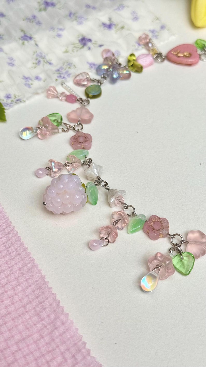 May include: A delicate necklace featuring a variety of pastel-colored beads and charms. The necklace includes pink, green, and clear beads, along with flower and leaf-shaped charms. The design is connected by silver-colored chain links.