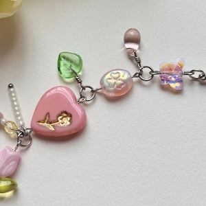 May include: A delicate necklace featuring a pink heart pendant with a gold flower design, complemented by iridescent beads, green leaf-shaped beads, and a butterfly charm. The necklace is crafted with silver-colored metal links and accents.