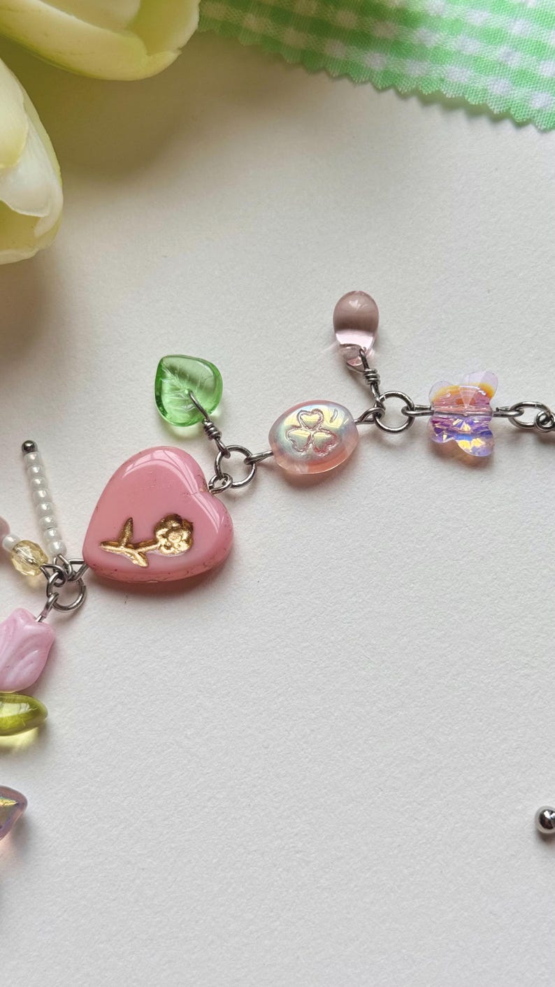 May include: A delicate necklace featuring a pink heart pendant with a gold flower design, complemented by iridescent beads, green leaf-shaped beads, and a butterfly charm. The necklace is crafted with silver-colored metal links and accents.