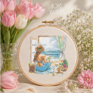 Puede incluir: Un bastidor de bordado de punto de cruz con una escena costera. La obra de arte representa a una persona con pelo largo y sombrero, sentada junto a una ventana abierta, mirando al mar. Un gato está cerca. El bordado está en un bastidor de madera, con el texto "RORY WOODS".