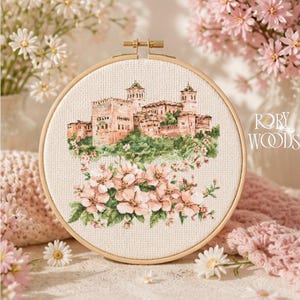 Puede incluir: Un bastidor de bordado de punto de cruz con una detallada representación de un castillo en una colina, rodeado de flores rosas. El bordado está sobre tela color crema, enmarcado por un aro de madera. El texto "RORY WOODS" es visible.