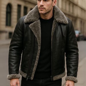 Op de afbeelding: Zwarte leren jas met een schapenvachtkraag en voering. De jas heeft een ritssluiting en een klassieke pilotenstijl. De schapenvacht is lichtgrijs en de jas wordt door een persoon gedragen.