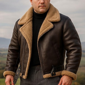 Puede incluir: Chaqueta de cuero marrón oscuro con cuello y forro de borrego. La chaqueta tiene cierre de cremallera, cinturón con hebilla y puños de borrego. El modelo lleva un cuello alto negro y pantalones grises. Paisaje de fondo.