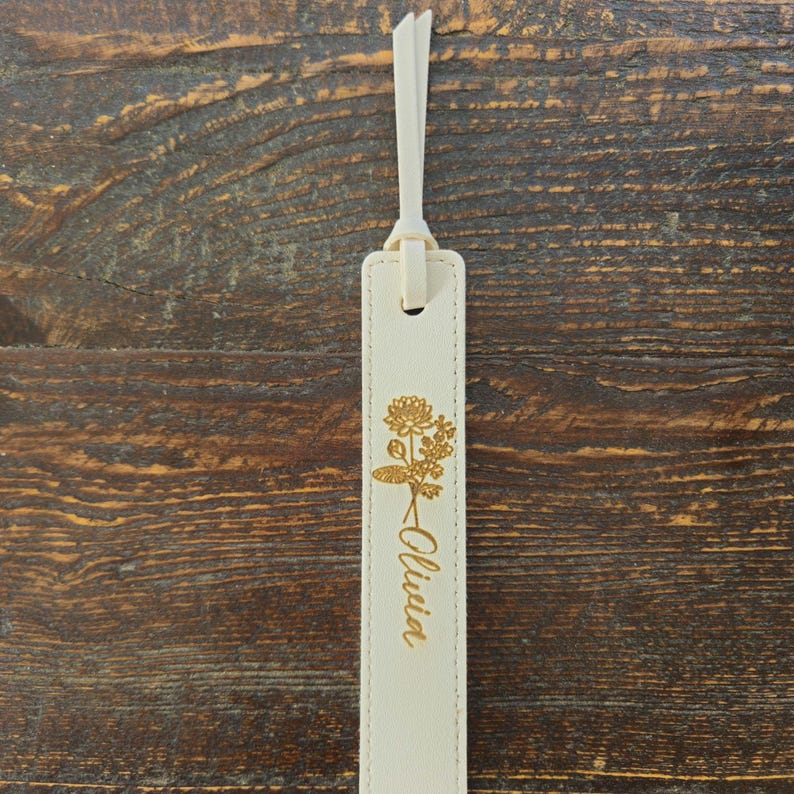 May include: Cream-colored leather bookmark with a gold floral design and the name "Olivia" in script. The bookmark has a matching cream-colored loop at the top for easy placement in a book. The background is a dark wood surface.