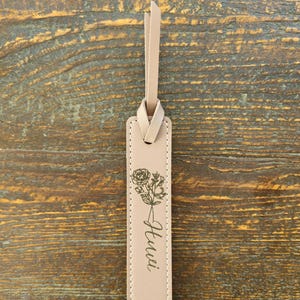 May include: A beige leather bookmark with a floral design and the word "Hui" printed in cursive. The bookmark has a loop at the top for easy placement in a book. The background is a wooden surface.