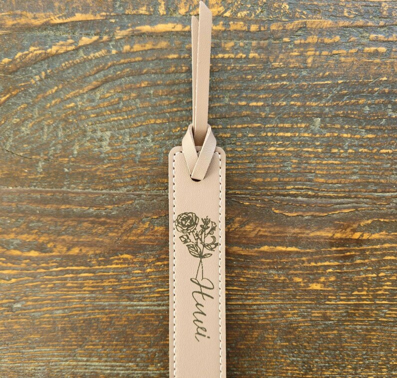 May include: A beige leather bookmark with a floral design and the word "Hui" printed in cursive. The bookmark has a loop at the top for easy placement in a book. The background is a wooden surface.