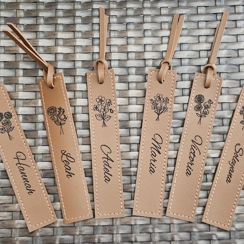 May include: Tan leather bookmarks with floral designs and names like Hannah, Leah, and Adela. Each bookmark has a stitched border and a matching leather loop. The background is a woven, light brown surface.