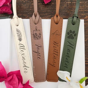 May include: Four leather bookmarks in cream, taupe, brown, and olive green. Each bookmark features a unique design: a feather, a candle with flowers, a mountain, and a paw print. Names are inscribed on each bookmark.