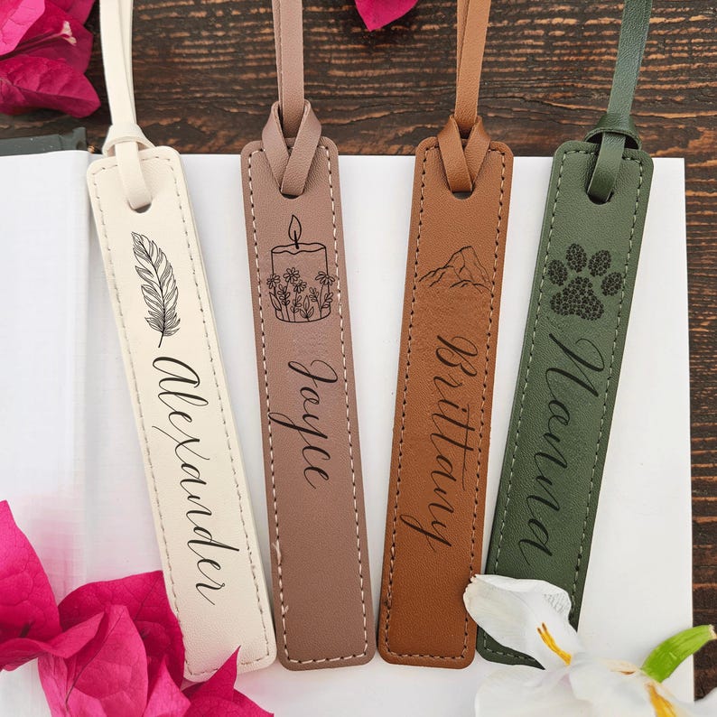 May include: Four leather bookmarks in cream, taupe, brown, and olive green. Each bookmark features a unique design: a feather, a candle with flowers, a mountain, and a paw print. Names are engraved on each bookmark: Alexander, Jayce, Brittany, and Nana.