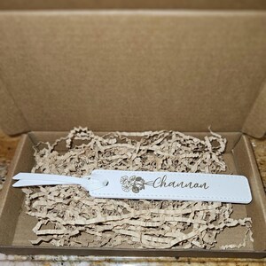 May include: A white leather luggage tag with the name "Channon" and a floral design, inside a cardboard box with brown paper filler. The tag has a loop for attachment.