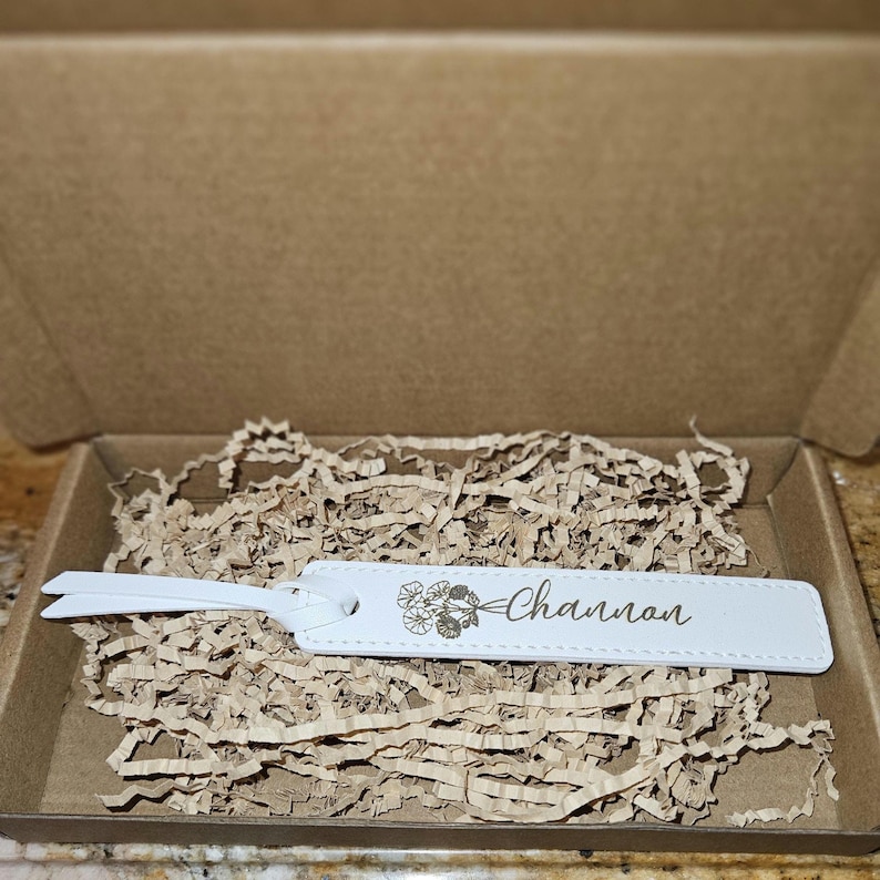 May include: A white leather luggage tag with the name "Channon" and a floral design, inside a cardboard box with brown paper filler. The tag has a loop for attachment.