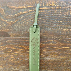 May include: A sage green leather bookmark with a floral design and the name "Charlotte" engraved. The bookmark has a loop at the top and a stitched border. The background is a wooden surface.