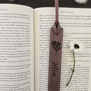 May include: A mauve leather bookmark with a floral design and the name "Mia" in cursive. The bookmark is rectangular and hangs over the pages of a book, with the text of the book visible in the background.