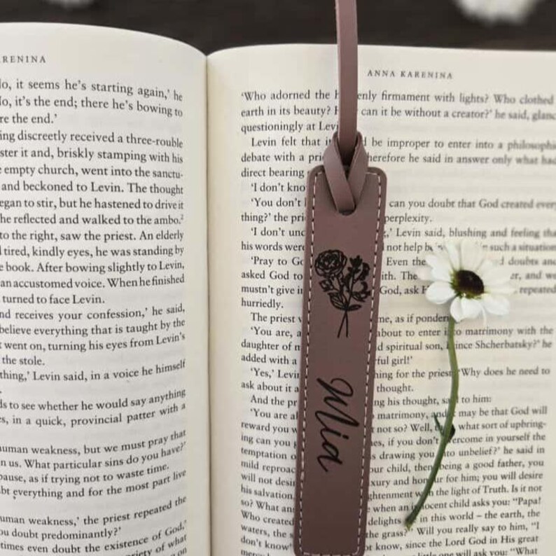 May include: A mauve leather bookmark with a floral design and the name "Mia" in cursive. The bookmark is rectangular and hangs over the pages of a book, with the text of the book visible in the background.
