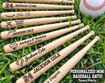 May include: A collection of light-coloured wooden mini baseball bats with various laser-engraved names, numbers, and baseball-themed graphics, accompanied by a white baseball with red stitching, all arranged on a patch of green grass.