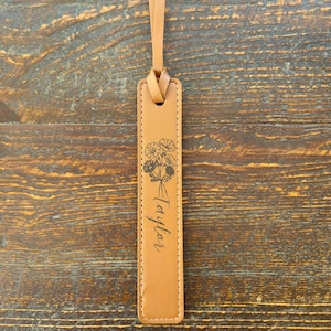 May include: A tan leather bookmark with a floral design and the name "Taylor" printed in cursive. The bookmark has a loop at the top for easy placement in a book. The background is a wooden surface.