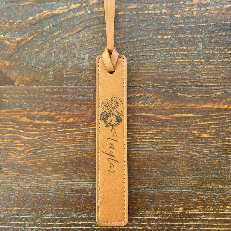 May include: A tan leather bookmark with a floral design and the name "Taylor" printed in cursive. The bookmark has a loop at the top for easy placement in a book. The background is a wooden surface.