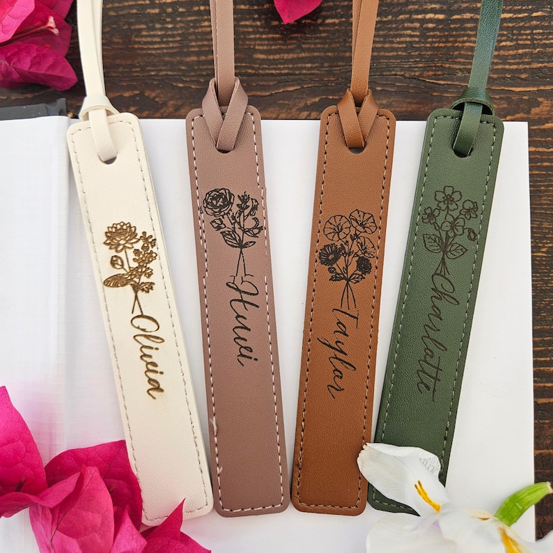 May include: Four personalized leather bookmarks in cream, mauve, brown, and olive green. Each bookmark features a floral design and a name: Olivia, Huwei, Taylor, and Charlotte. The bookmarks are displayed on a wooden surface.