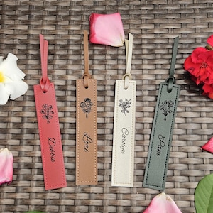 May include: Four leather bookmarks in shades of pink, tan, cream, and gray, each with a floral design and a name: Debbie, Lori, Caroline, and Pam. Each bookmark has a loop at the top and is decorated with a stitched border.