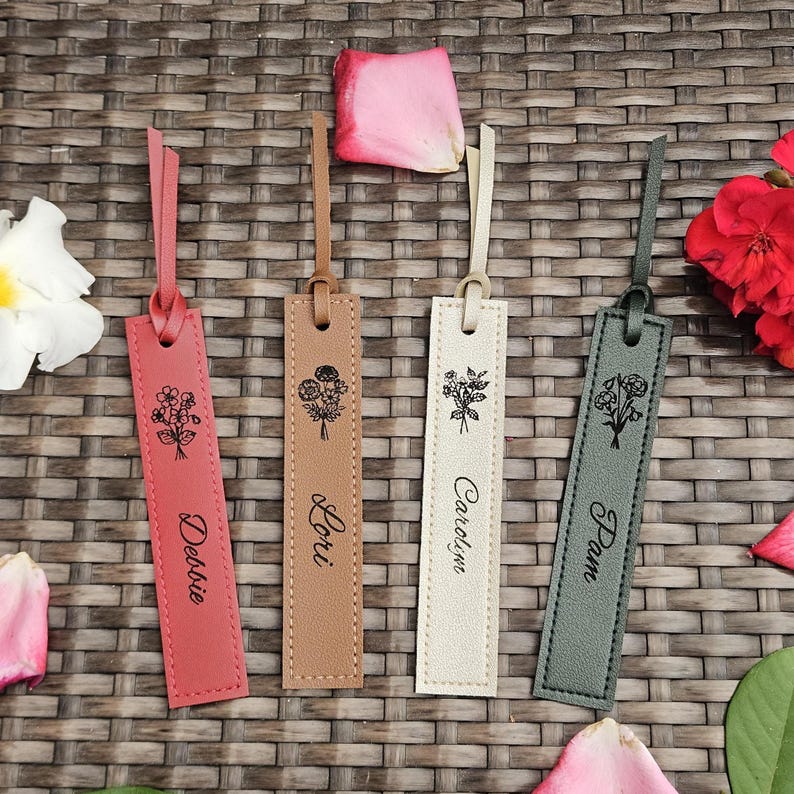 May include: Four leather bookmarks in shades of pink, tan, cream, and gray, each with a floral design and a name: Debbie, Lori, Caroline, and Pam. Each bookmark has a loop at the top and is decorated with a stitched border.