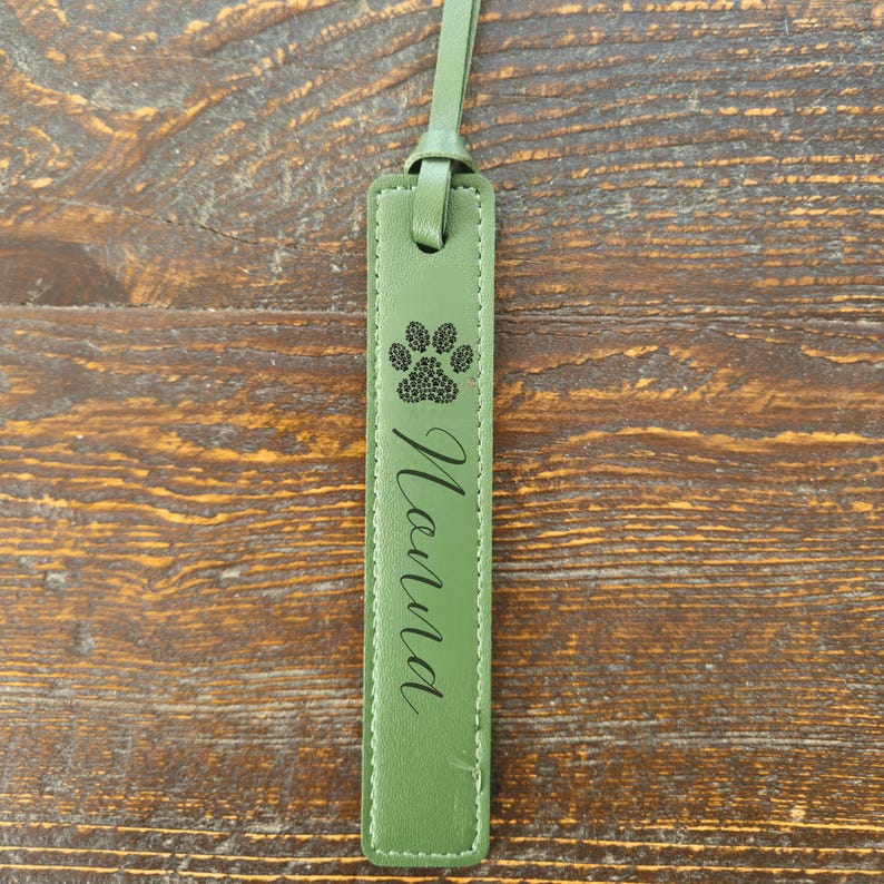 May include: Olive green leather bookmark with a paw print design and the name "Nanna" in cursive script. The bookmark has a loop at the top for easy placement in a book. The stitching is white.