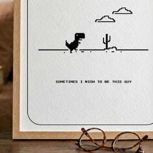 Può includere: Una stampa incorniciata con un dinosauro nero pixelato, un cactus e nuvole su sfondo bianco. Il testo "SOMETIMES I WISH TO BE THIS GUY" è in basso. Un paio di occhiali rotondi poggiano su una superficie di legno.