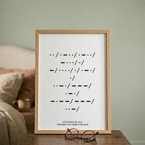 Può includere: Una stampa incorniciata con una cornice in legno chiaro mostra il codice Morse in nero su sfondo bianco. Il codice Morse compone un messaggio, con il testo "(I'll be there for you)" in basso. Un paio di occhiali rotondi poggiano sotto la cornice.