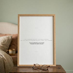 May include: A framed print with a light wood frame, displaying a line of Morse code and the text "(If you're reading this, you're an idiot) Translation not visible in the print." Round eyeglasses rest on a wooden table.