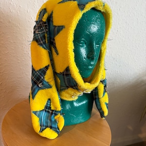 Pode incluir: Um capuz de tecido felpudo amarelo brilhante com remendos em forma de estrela em xadrez azul e verde. O capuz inclui painéis laterais longos e é decorado com vários alfinetes de segurança de metal prateado fixados por todo o material.