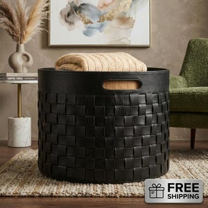 Puede incluir: Una cesta de almacenamiento redonda de color negro con un patrón exterior trenzado y asas laterales integradas. La cesta está cubierta por una manta de punto beige texturizada y doblada, creando un contraste entre la estructura oscura y la tela.