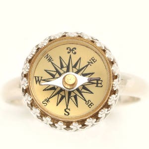 May include: A silver ring featuring a compass face. The compass face is gold with black markings and a black needle pointing north. The ring is a unique and stylish accessory.