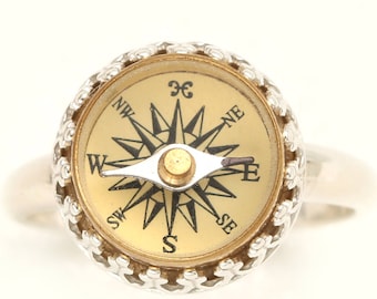 May include: A silver ring with a compass face. The compass face is gold with black markings and a black needle pointing north. The ring is a unique and stylish accessory.
