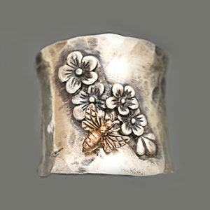 Puede incluir: Un anillo de plata con una superficie texturizada y un dise&ntilde;o de flores y una abeja. La abeja es de oro.