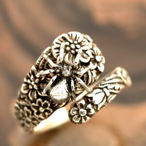 May include: A silver ring with a detailed design of a spider and flowers. The spider is in the center of the ring and has a small clear stone in its body.