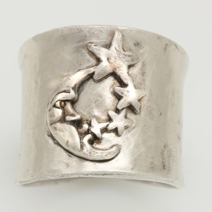 May include: A silver ring featuring a crescent moon and stars design. The ring is wide and has a hammered finish.
