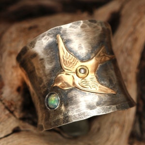 May include: A silver cuff bracelet with a hammered finish features a gold-toned bird design with an opal gemstone. The bird has a pearl-like accent in the center of its chest.