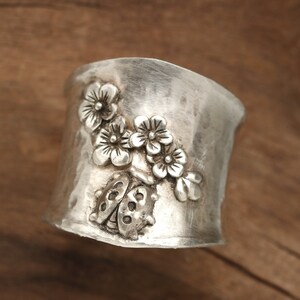 May include: A silver ring with a textured finish features a design of a ladybug and flowers. The ring is on a wooden surface.