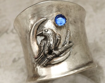 May include: A silver ring featuring a blue gemstone and an intricate design of a raven perched on a branch. The ring is wide and has a textured finish.
