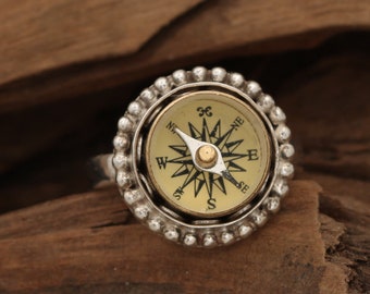 May include: A silver compass ring with a gold face and black markings. The compass face has the letters N, NE, E, SE, S, SW, W, and NW around the edge. The compass needle points to the north.