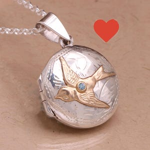 May include: A round silver locket pendant on a silver chain featuring an engraved floral pattern and a gold-toned swallow bird overlay. The bird has a small, light blue gemstone set in its center. A red heart icon is positioned above the locket.